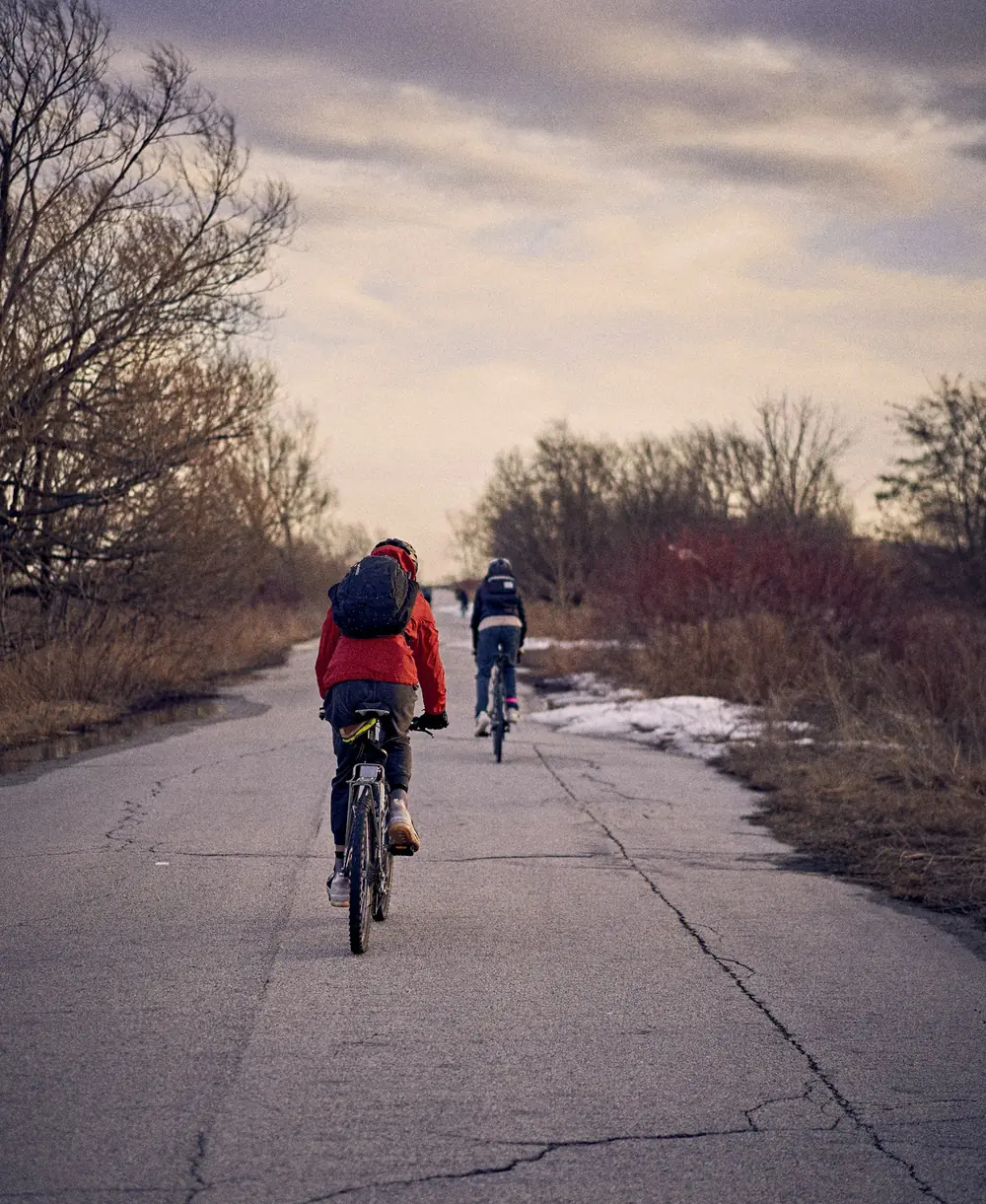 Two cyclists riding a paved path at Tommy Thompson Park under a cloudy evening sky